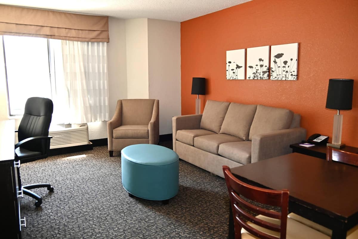 Habitación de hotel con un sofá color beige, un taburete azul, una mesa de madera y una silla de oficina negra. Hay dos lámparas de pared y una ventana con cortinas translúcidas.