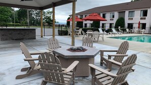 A covered outdoor seating area with Adirondack chairs and a stone fire table, overlooking a pool and a building with red umbrellas. - Sleep Inn & Suites Spring Lake - Fayetteville near Fort Bragg (Spring Lake)