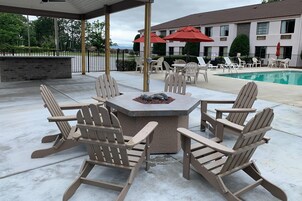 A covered outdoor seating area with Adirondack chairs and a stone fire table, overlooking a pool and a building with red umbrellas.