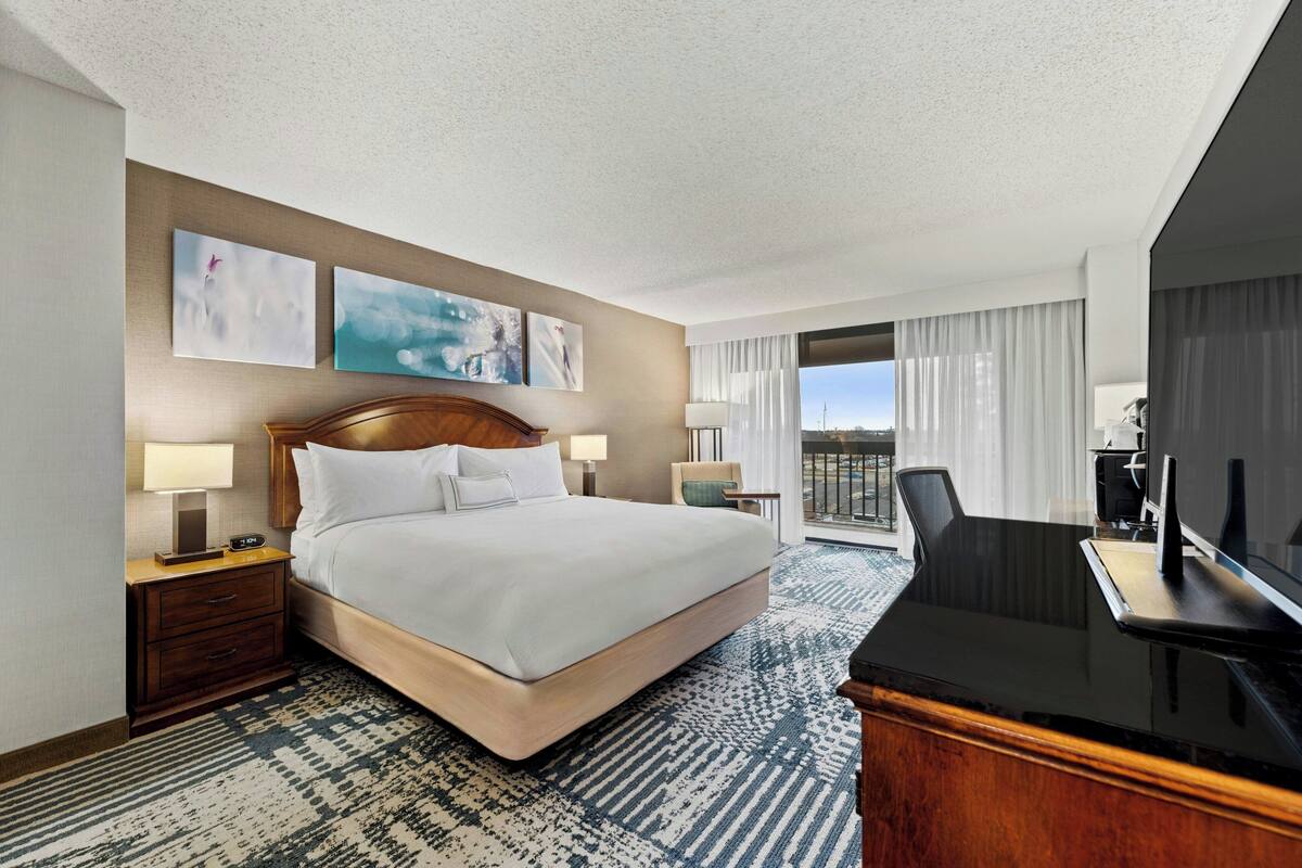 Habitación de hotel con una cama grande, un escritorio con televisor, una silla, una mesita de noche con lámpara y vistas al exterior por la ventana.