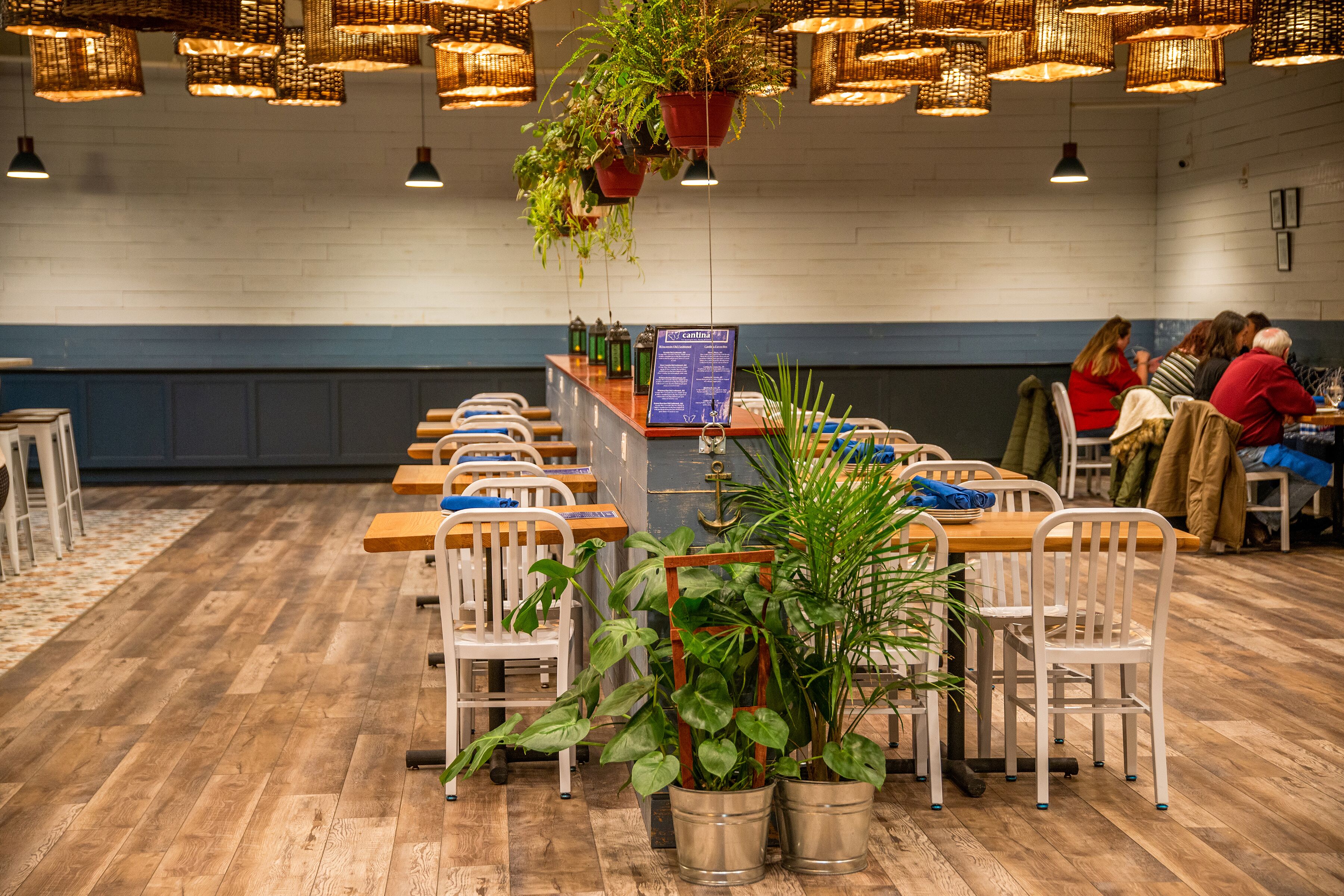 A restaurant with wooden tables and chairs, hanging plants, and a menu board.