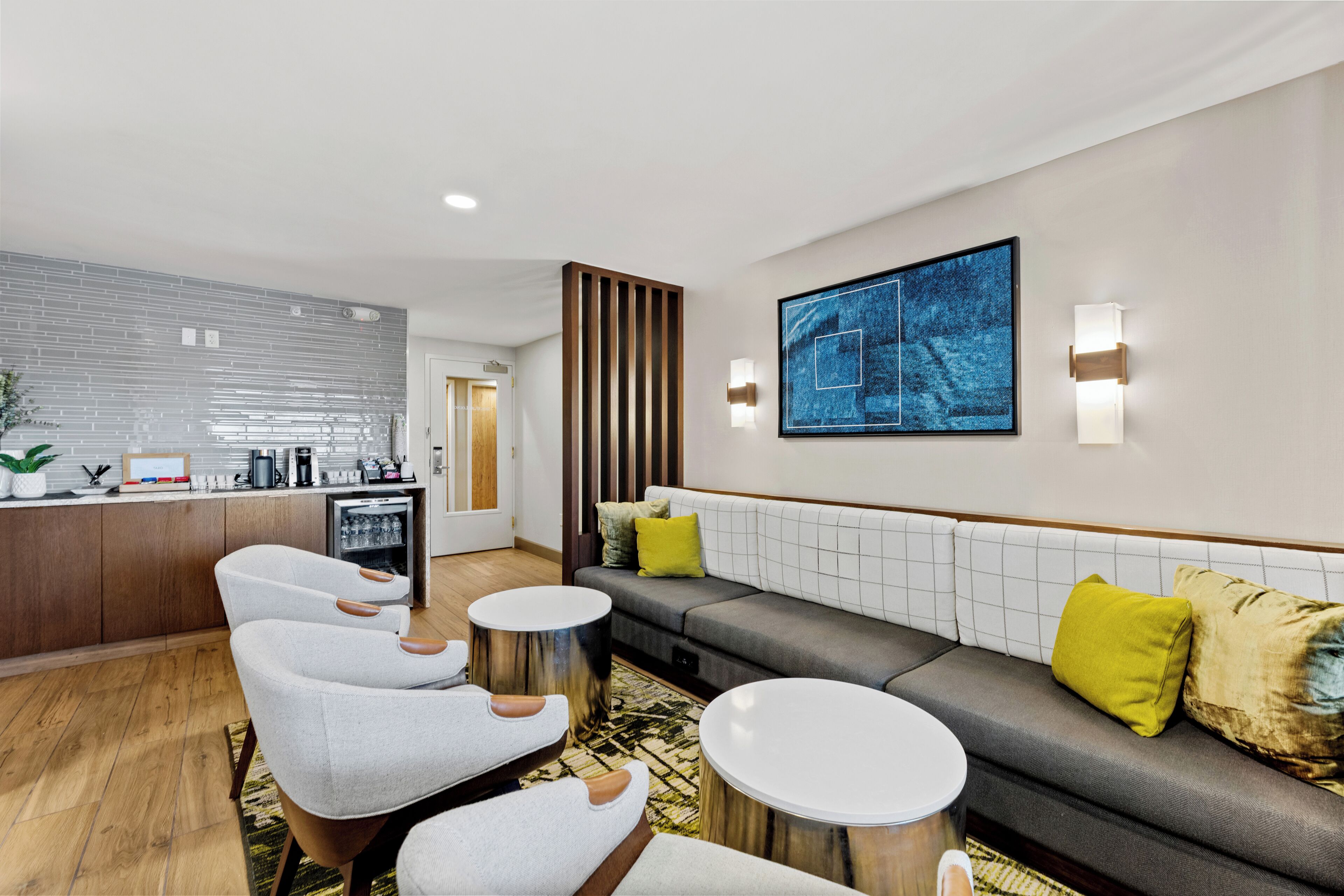 A modern hotel lobby with a seating area, a kitchenette, and a large abstract painting on the wall.