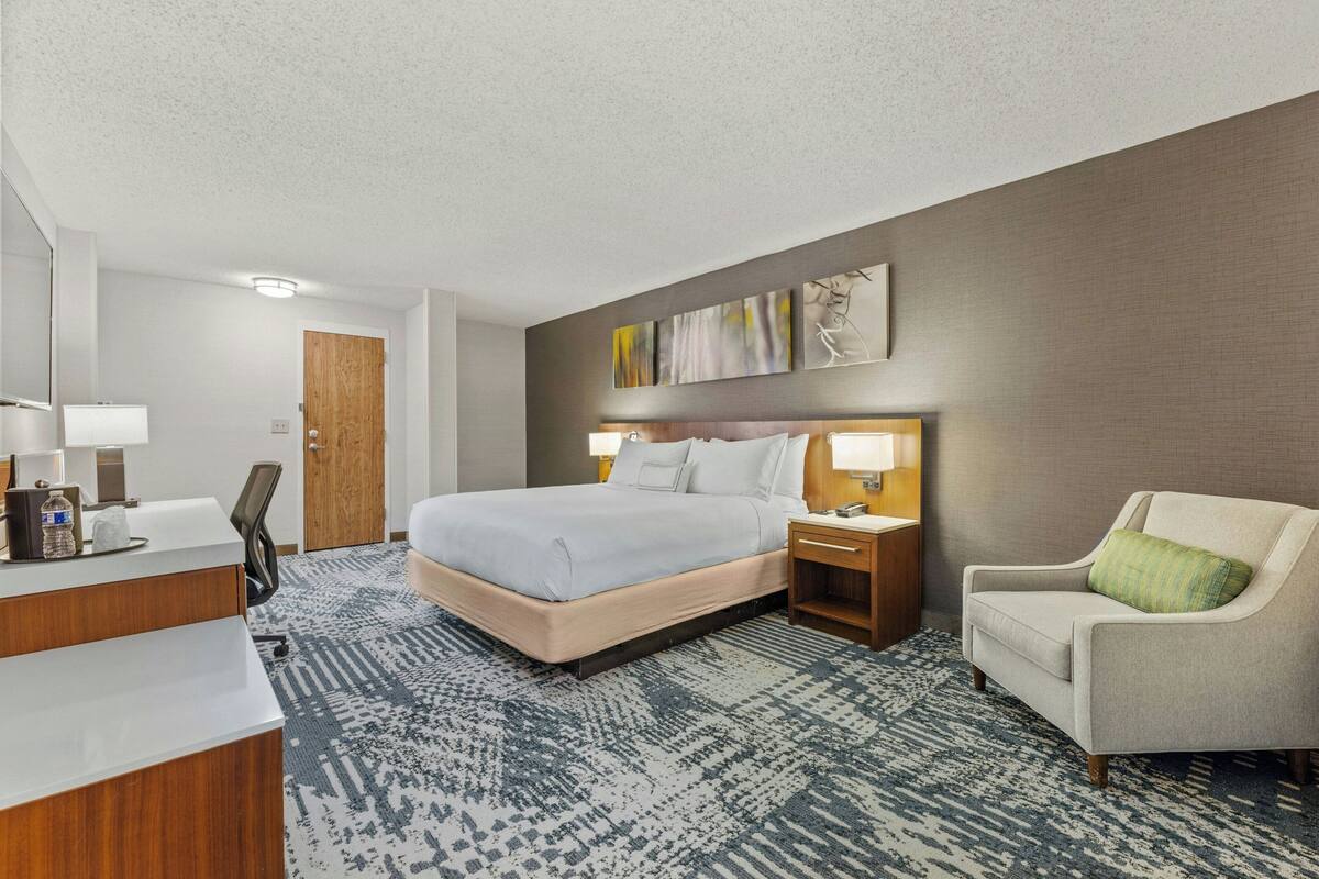 Una habitación de hotel con cama, mesitas de noche, una silla, un escritorio y un espejo.