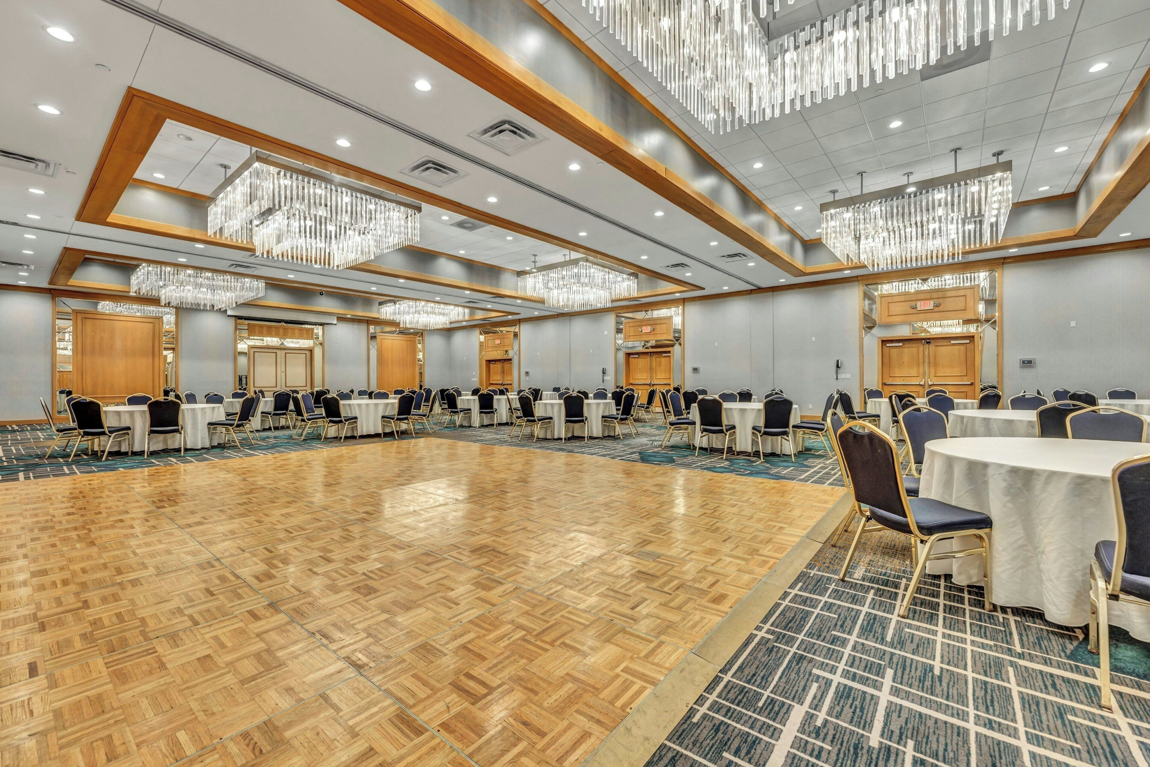 Un amplio salón de conferencias con múltiples mesas y sillas, suelo de madera y modernos candelabros.