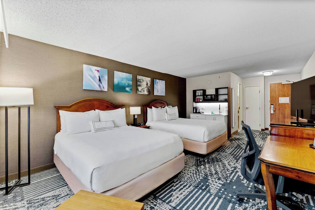 Habitación de hotel con dos camas, un escritorio, una silla, una lámpara y una obra de arte en la pared.