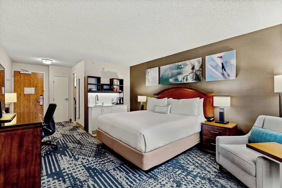 Habitación de hotel con una cama grande, un escritorio, una silla, una lámpara de escritorio, una mesita de noche y una pared con dos cuadros enmarcados.