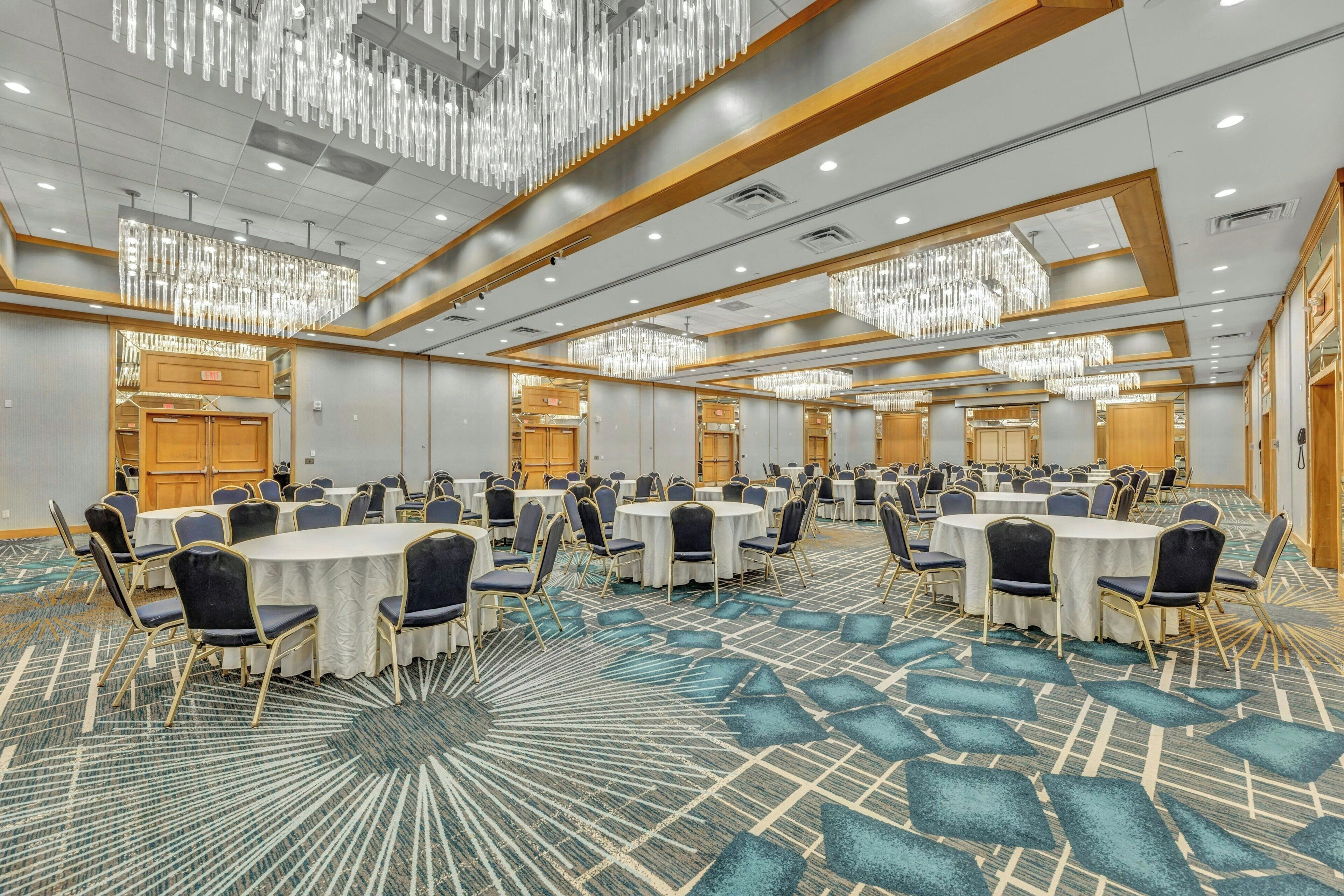 A large conference room with round tables and chairs, chandeliers, and a patterned carpet.
