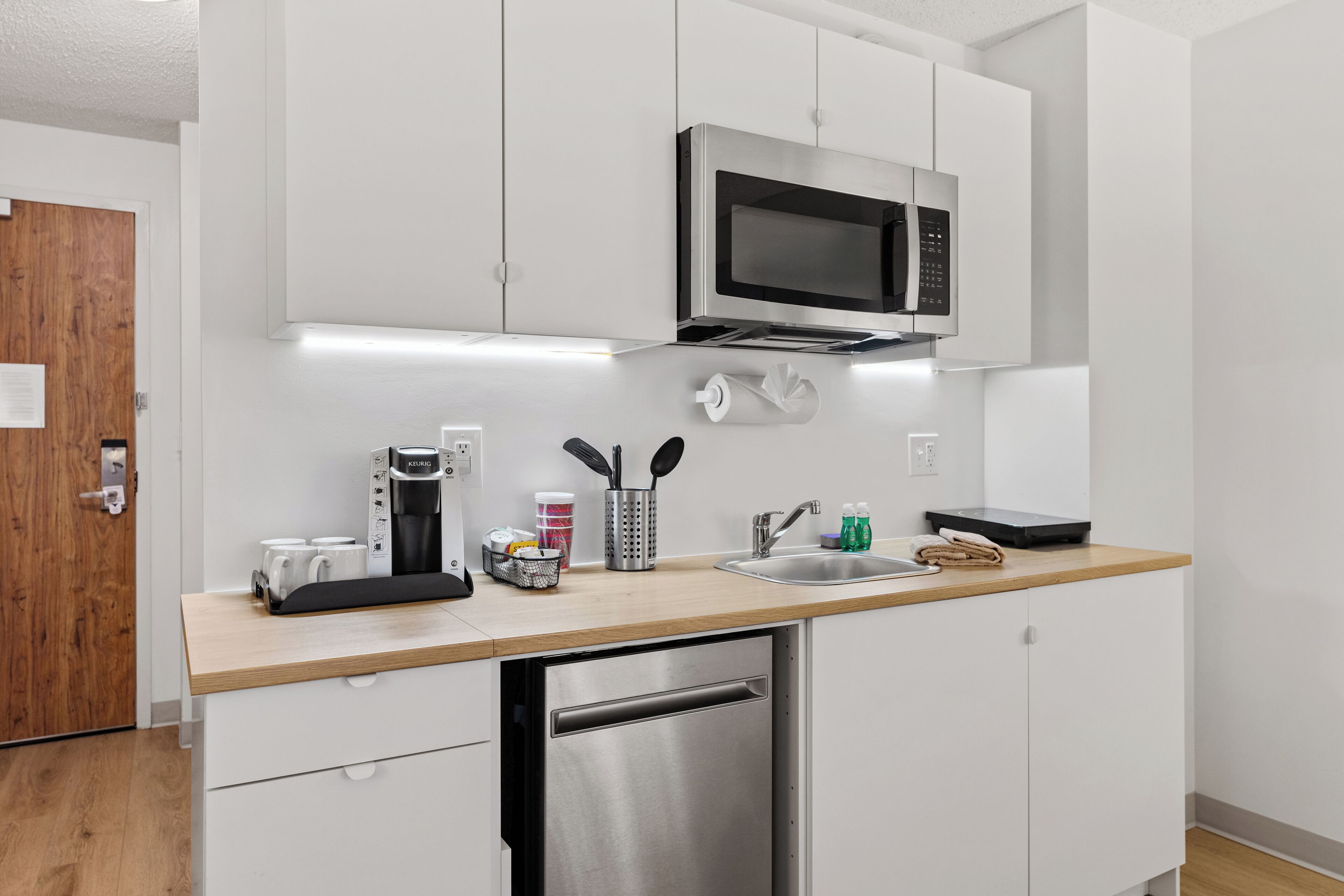 Una cocina moderna con armarios blancos, lavavajillas de acero inoxidable, microondas integrado, cafetera y un lavabo con grifo.