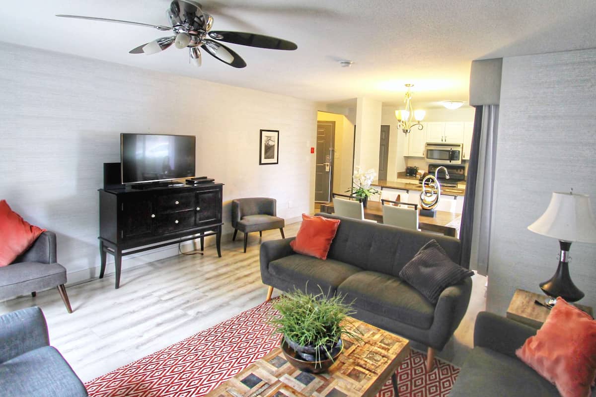 A living room with a sofa, armchairs, a TV, a ceiling fan, and a kitchen in the background.