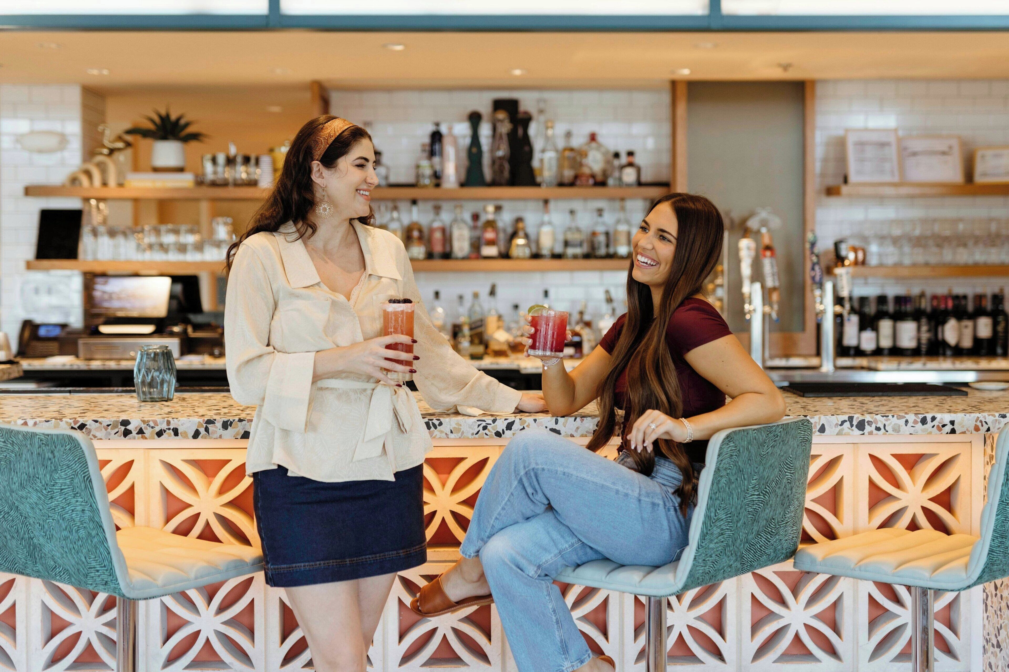 Two people seated at a bar with a patterned counter and shelves of bottles in the background.