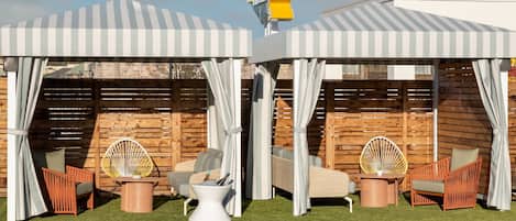 A pool area with two covered seating areas, each with a chair and a table, and a wooden fence in the background.
