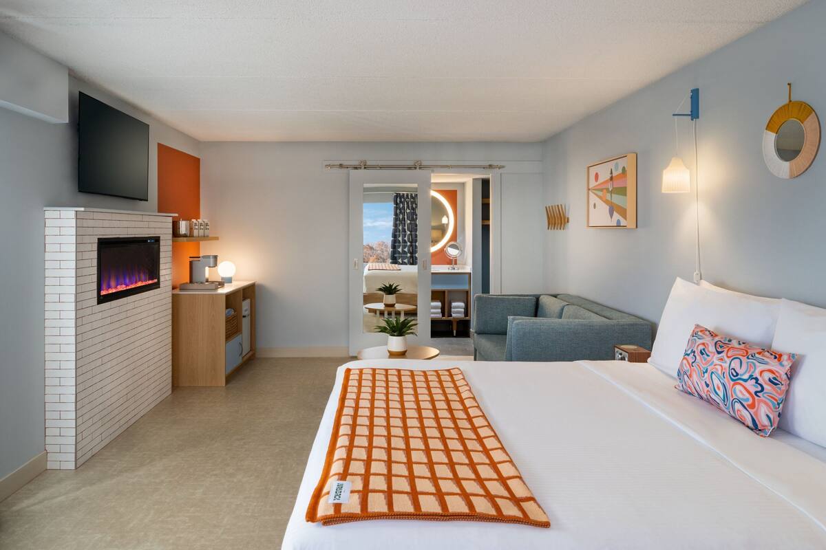 A modern hotel room with a fireplace, a bed with a patterned blanket, a sofa, and a desk with a lamp.