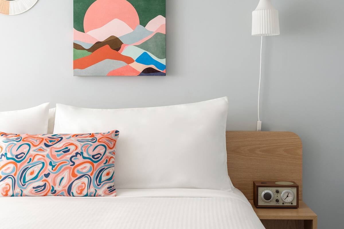 A modern bedroom with a bed, pillows, a wall-mounted painting, a bedside table with a radio, and a hanging lamp.