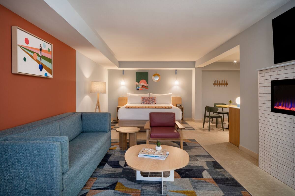 A modern hotel room with a sofa, a round coffee table, a bed with a headboard, a desk, and a fireplace.