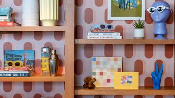 A room with a patterned wallpaper, wooden shelves, and various decorative items including books, a cocktail shaker, and a blue sculpture.