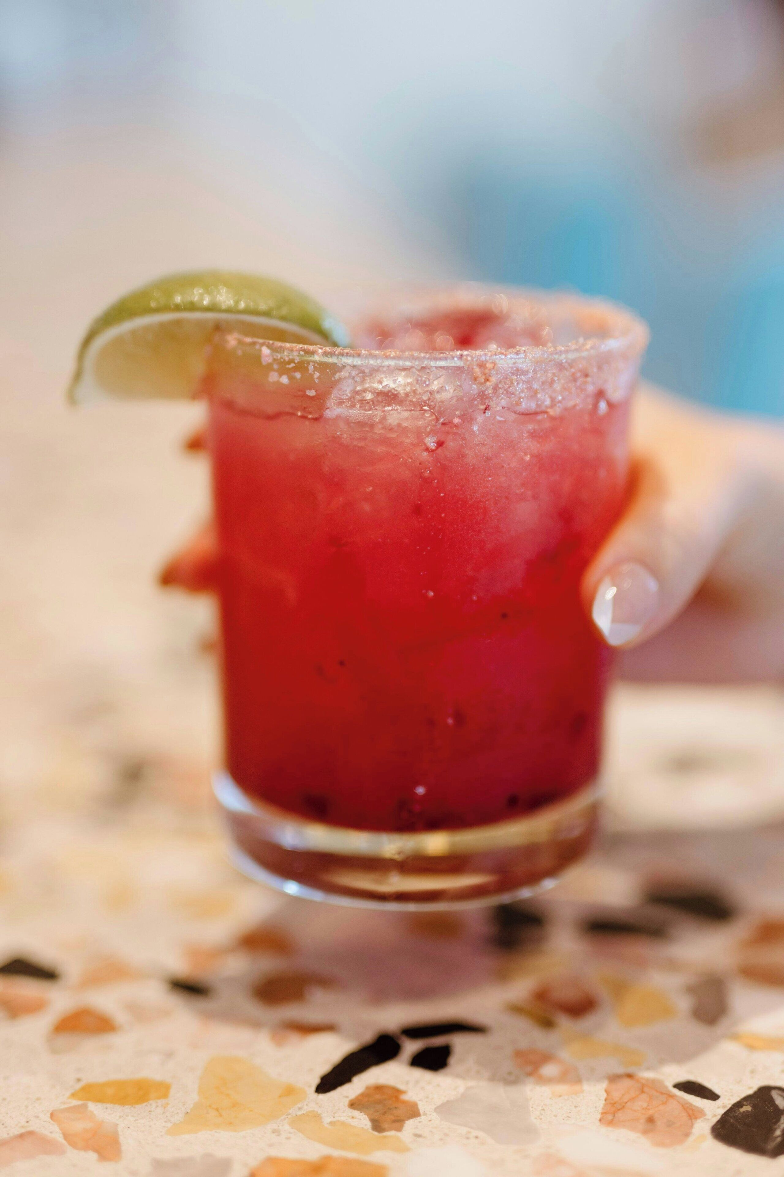 A glass of red beverage with a lime garnish, held by a hand.