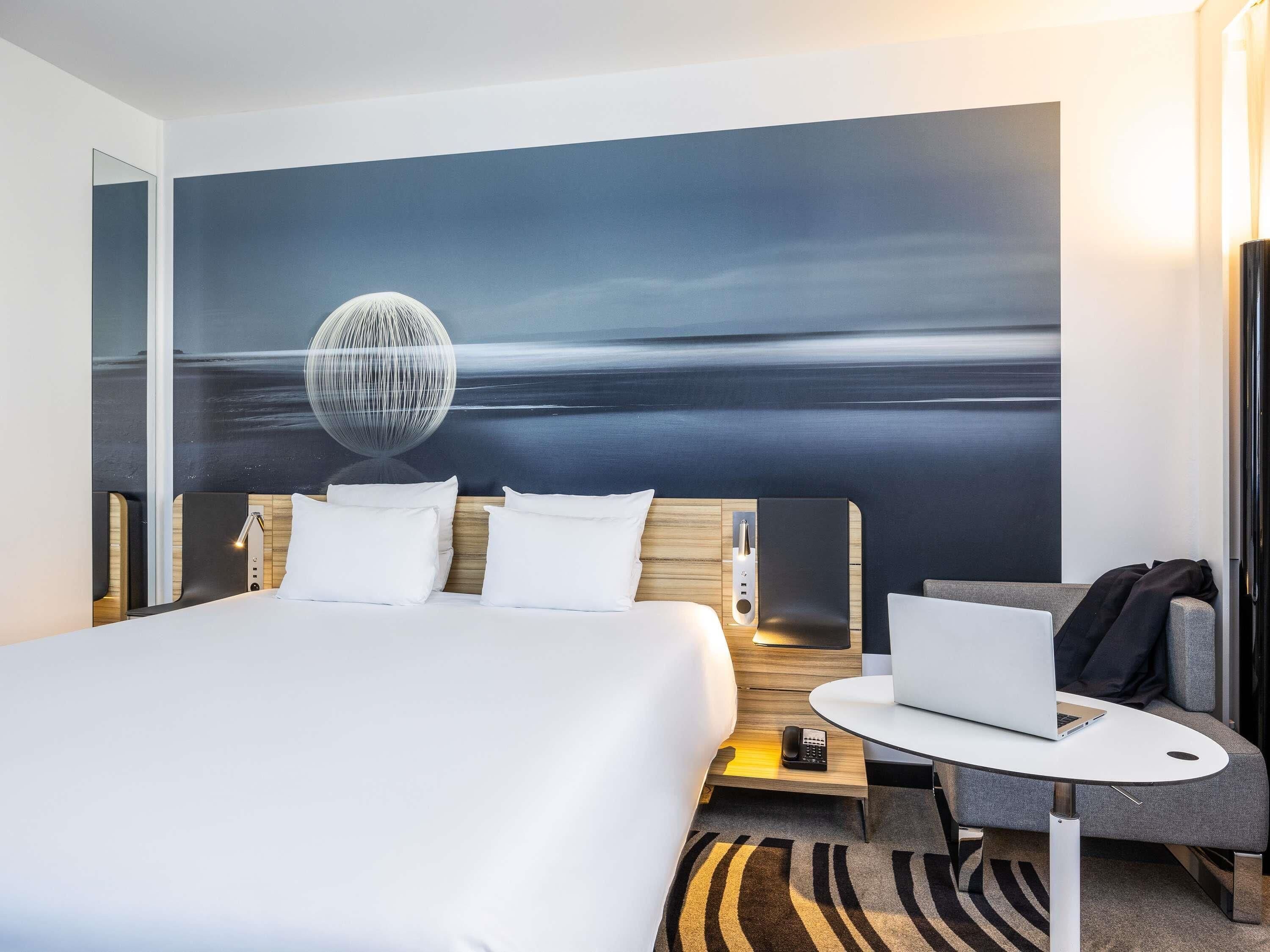 Habitación de hotel con una cama, un escritorio con una computadora portátil, un teléfono y un cuadro de un paisaje.