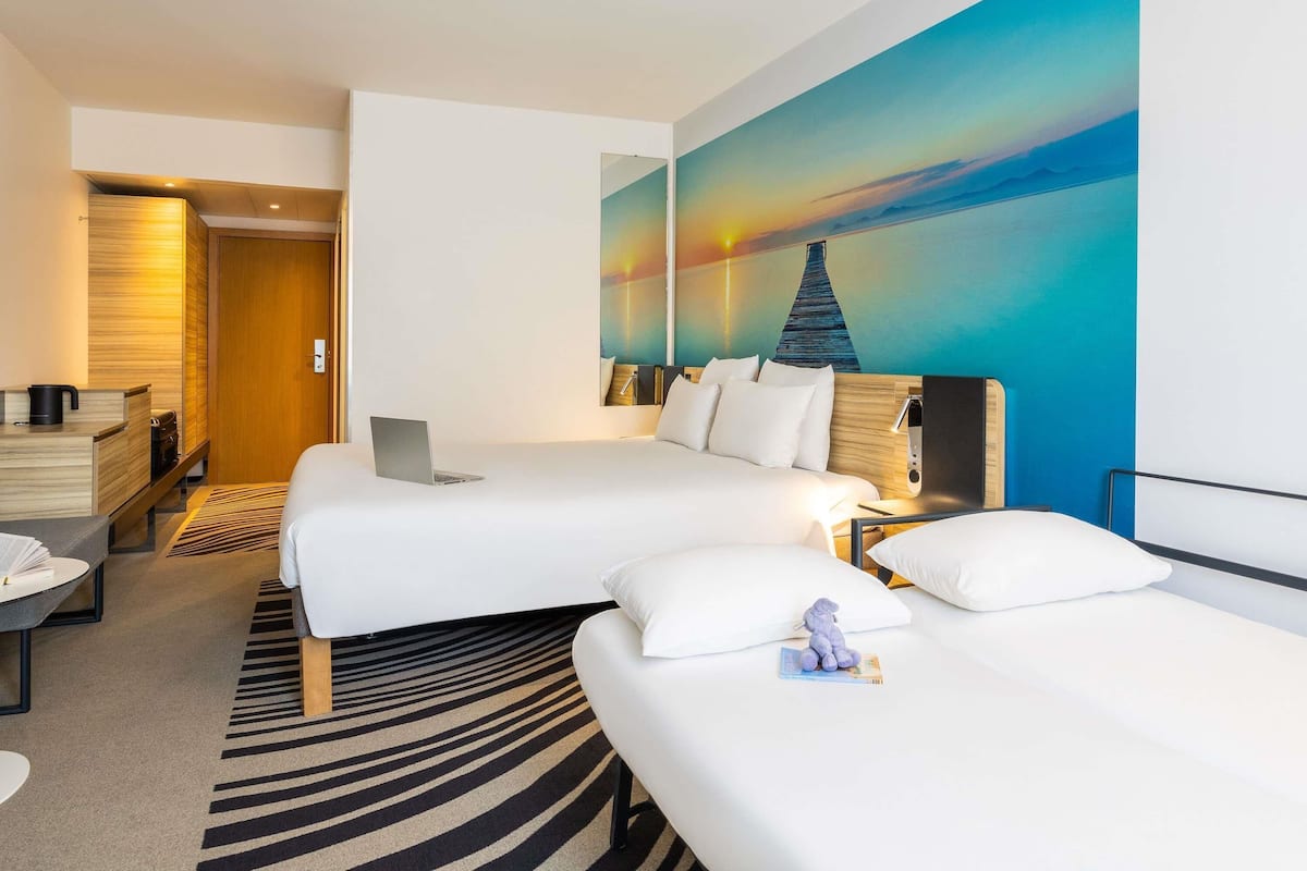Une chambre d’hôtel avec deux lits, un bureau, un ordinateur portable, une petite statue et un tableau représentant un coucher de soleil.