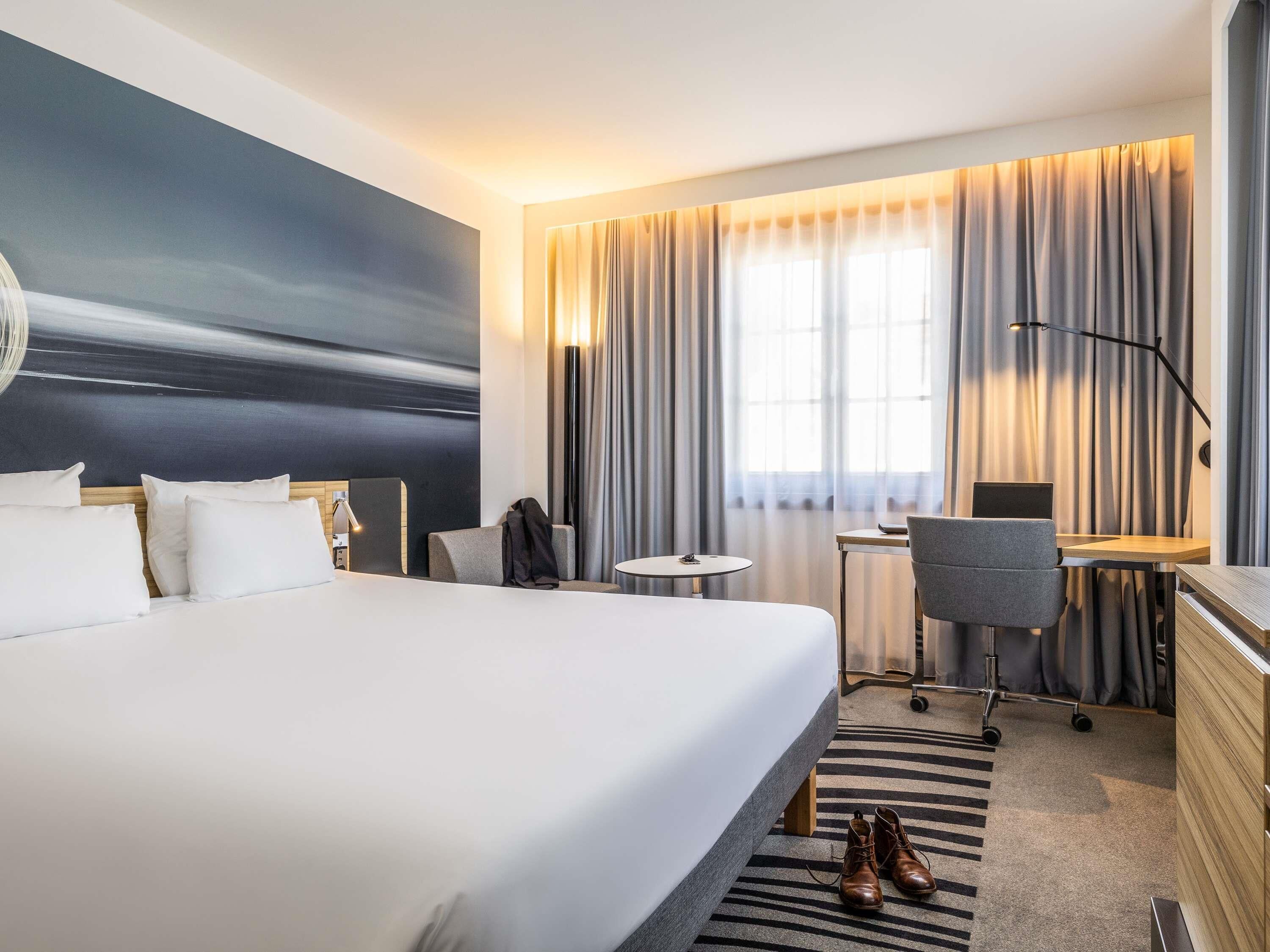 A hotel room with a bed, a desk, a chair, a small table, a coat rack, and a large window with curtains.
