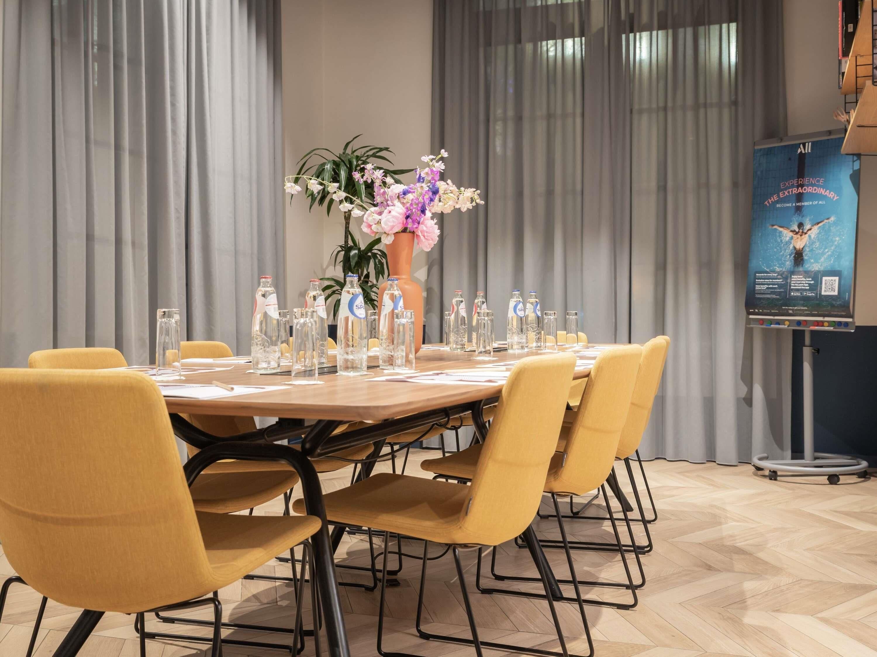 Un salón de conferencias con una mesa redonda, sillas amarillas, un centro de mesa floral, botellas de agua y un cartel en la pared.