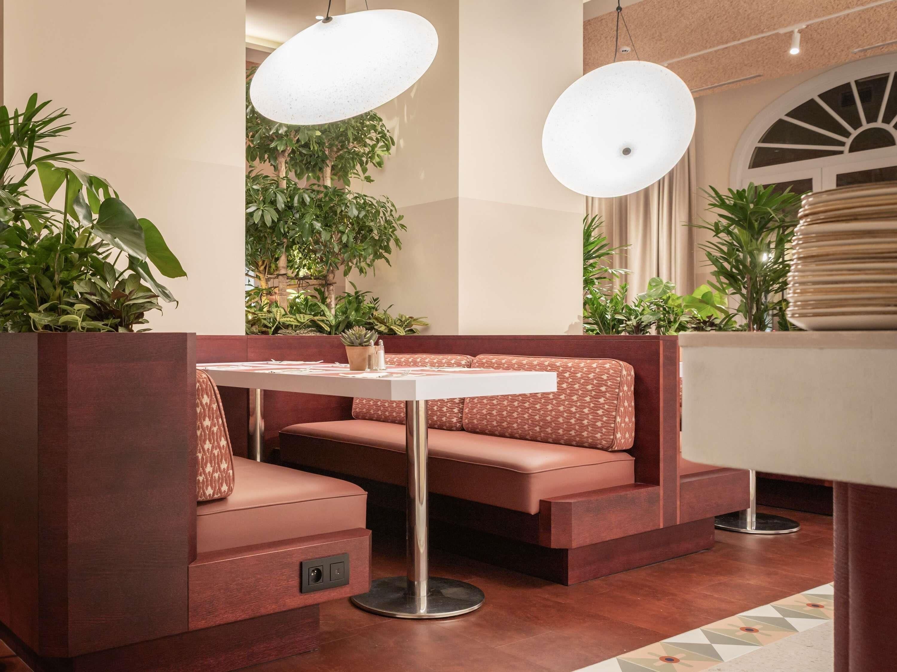 A modern interior with a seating area featuring a white table and a cushioned bench, surrounded by potted plants and pendant lighting.