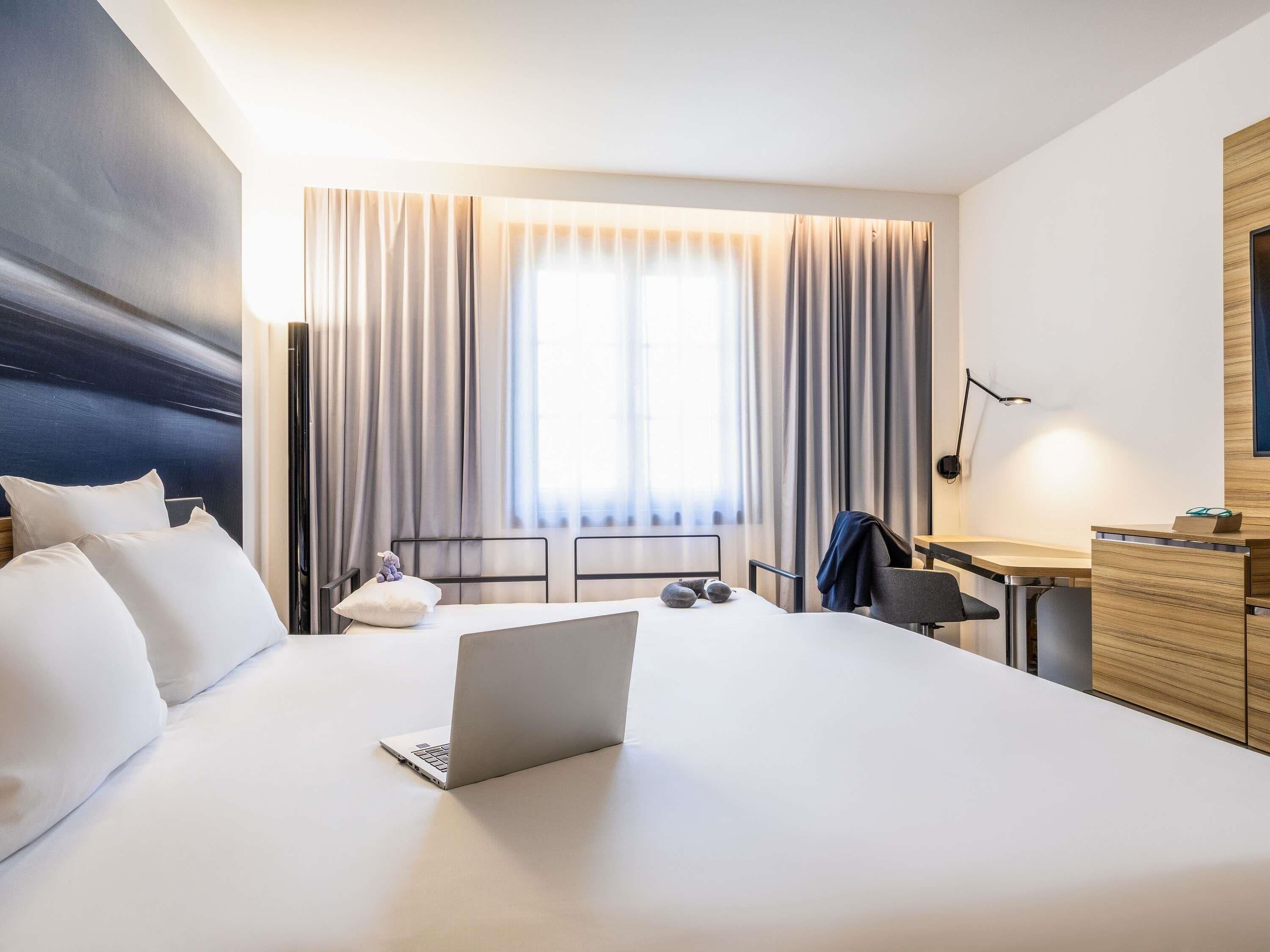 A hotel room with a bed, a desk, a chair, a lamp, and a window with curtains.