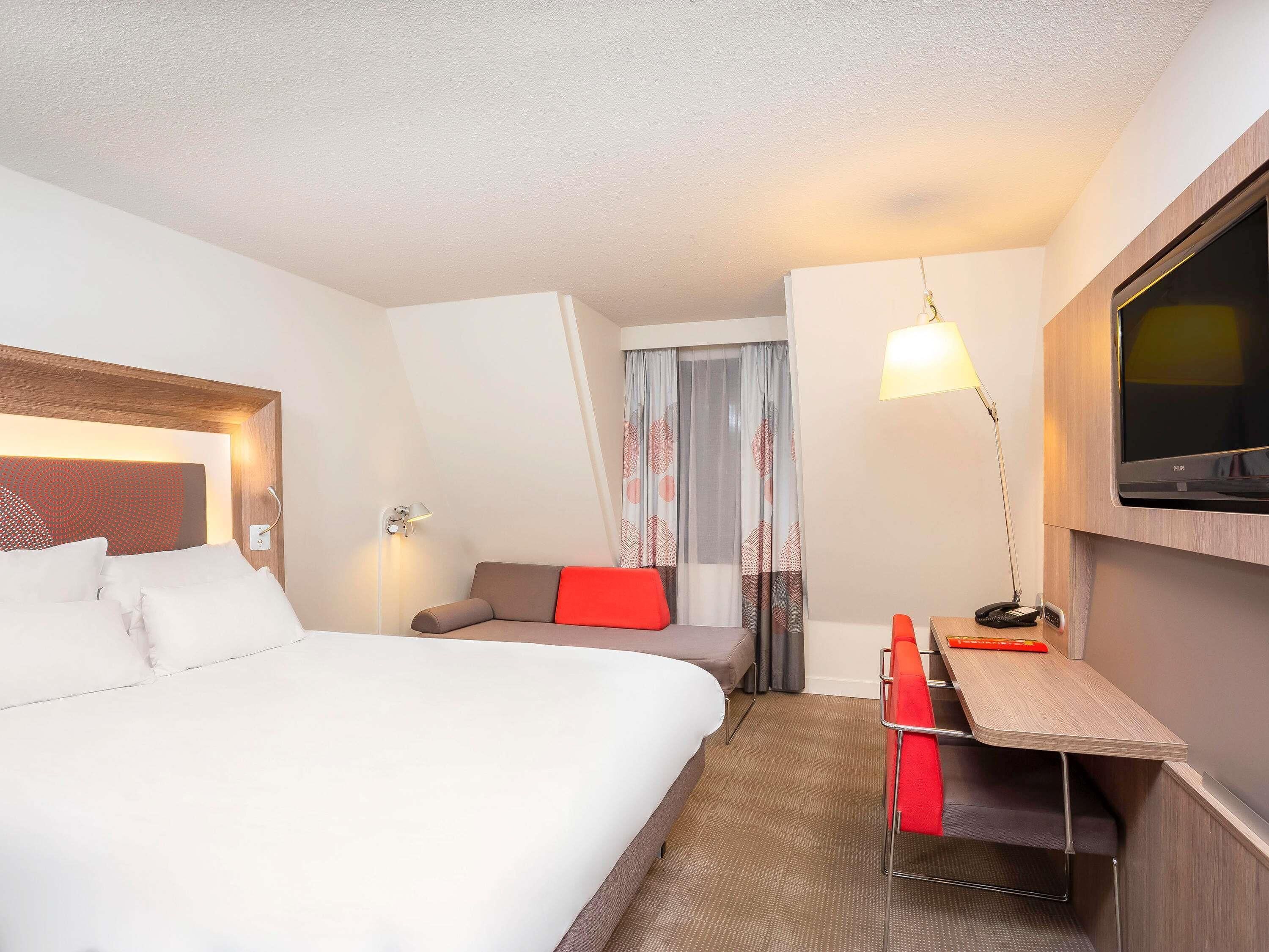 A hotel room with a bed, a desk with a chair, a television, and a window with curtains.
