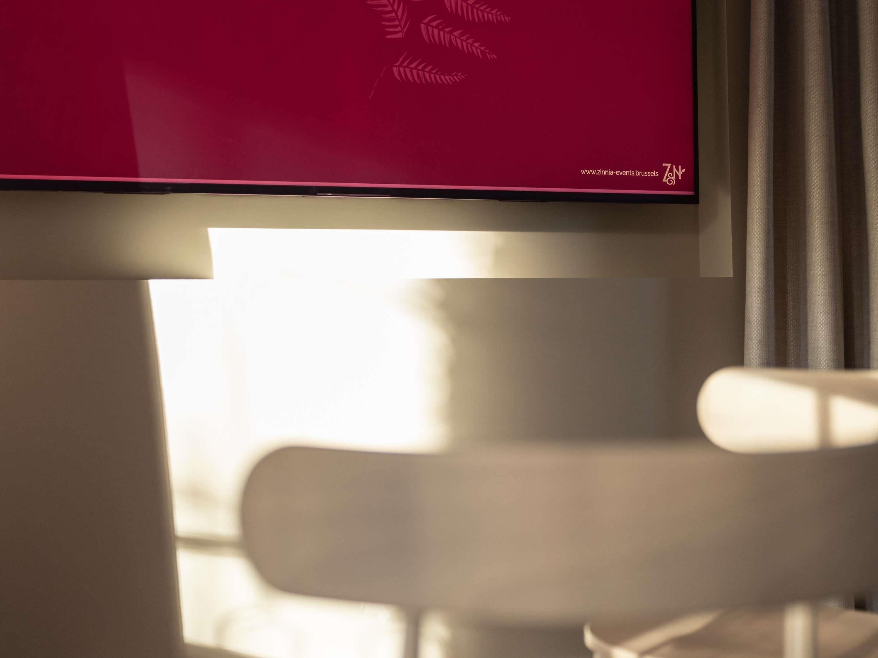 A room with a television mounted on the wall, a window with curtains, and a white chair.