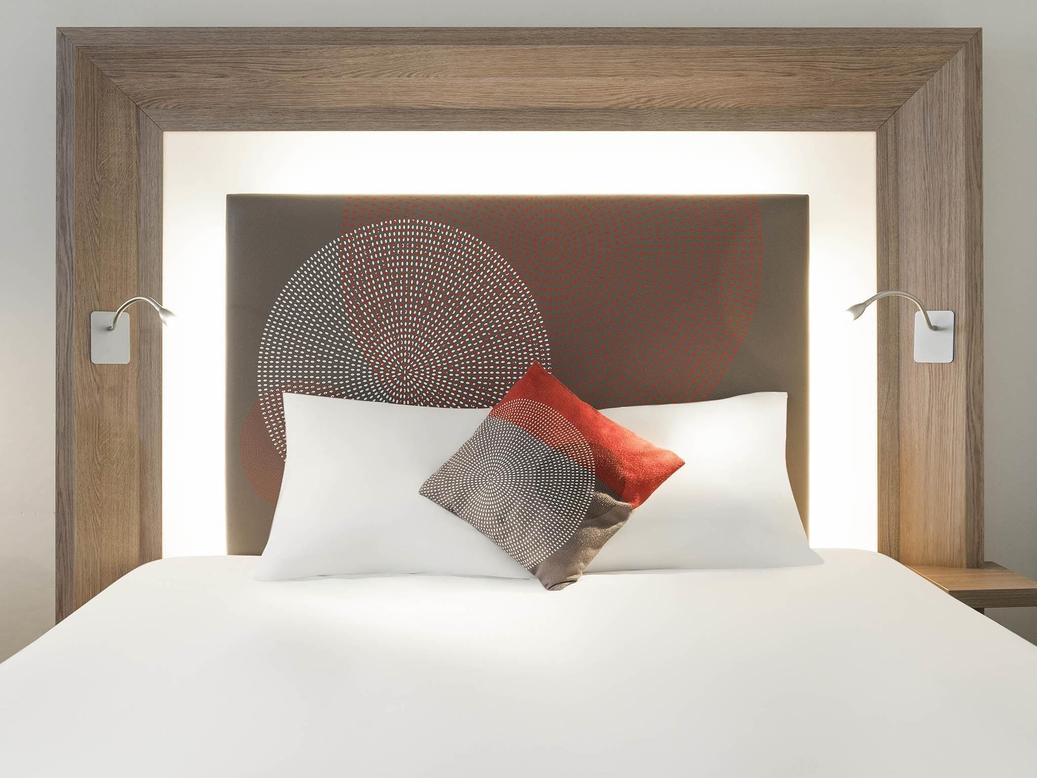 A neatly made bed with a wooden headboard, two wall-mounted light fixtures, and a large, circular, patterned pillow.