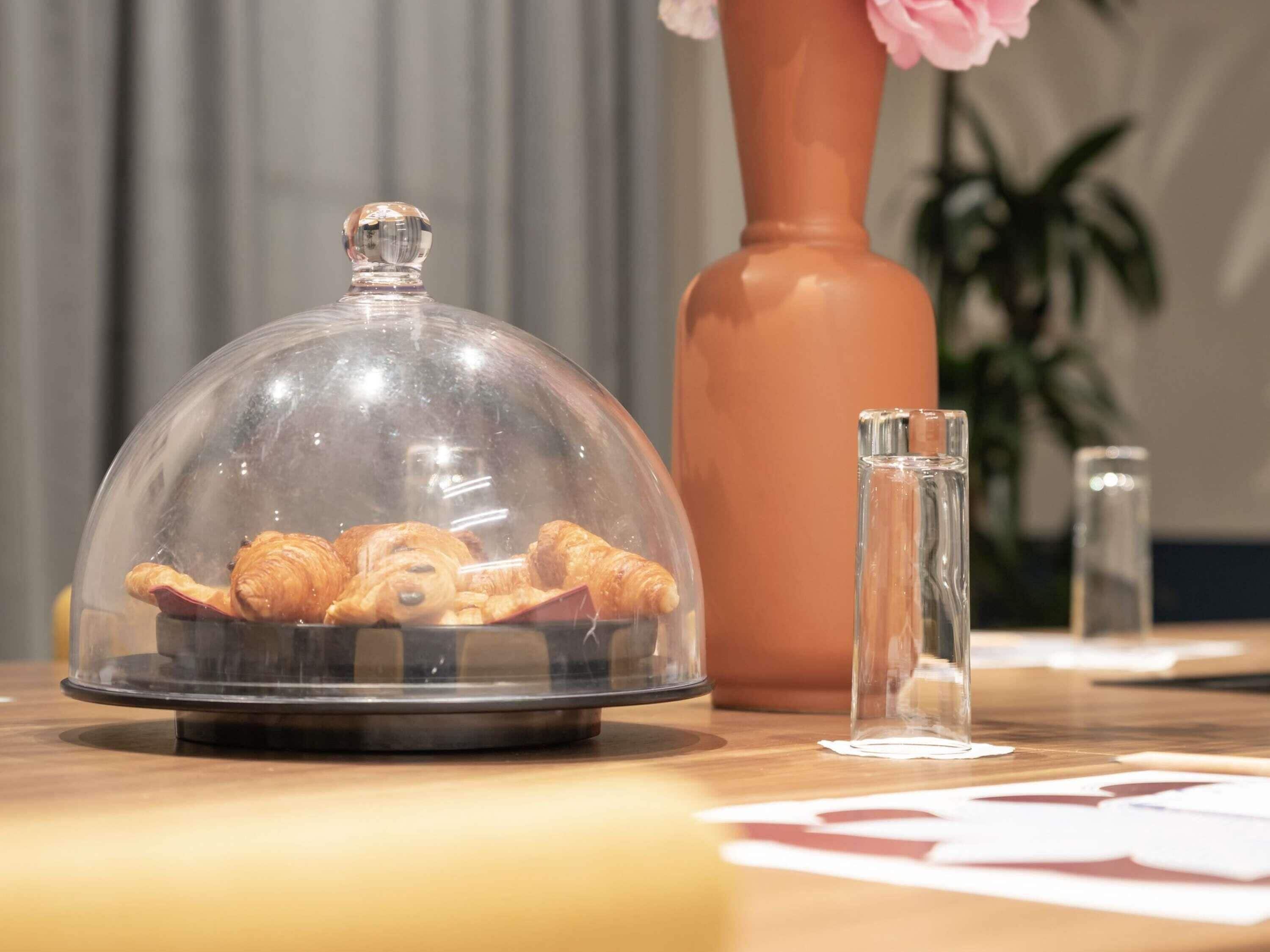 A table with a glass dome displaying pastries, a tall vase with pink flowers, and a clear glass bottle.