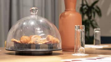 A table with a glass dome displaying pastries, a tall vase with pink flowers, and a clear glass bottle.