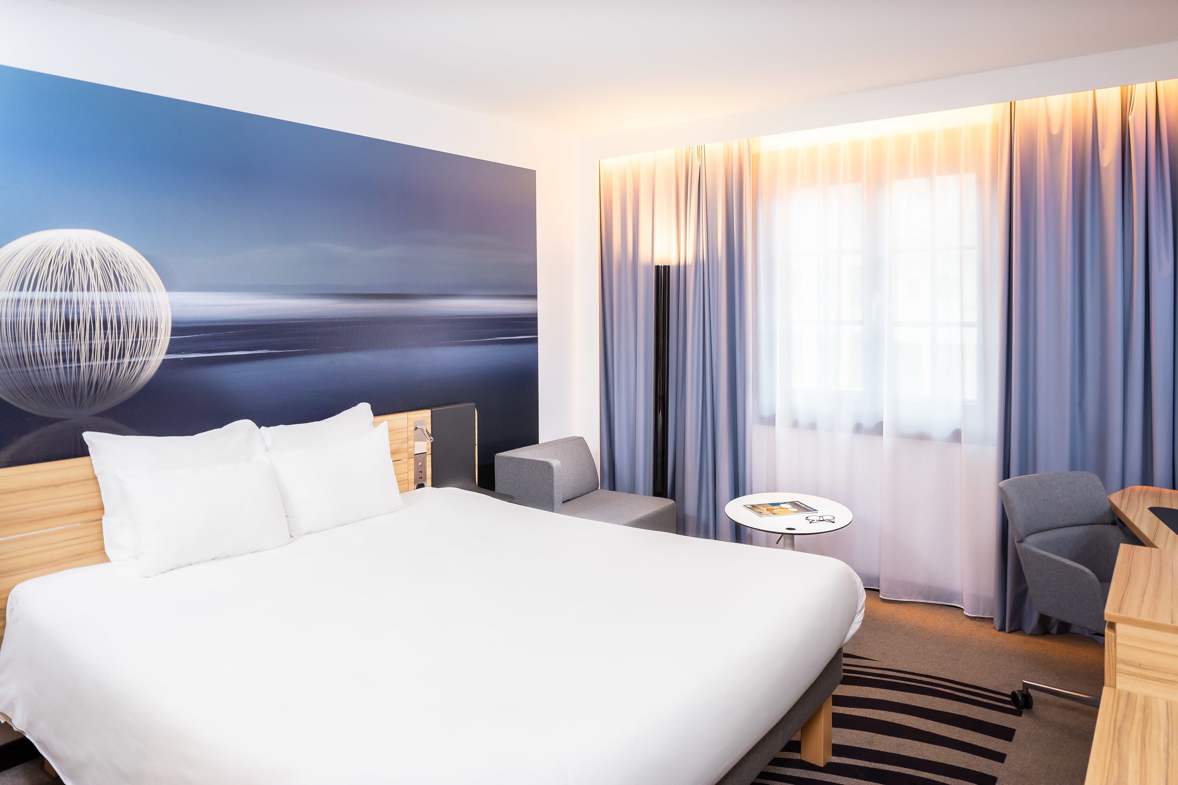 A modern hotel room with a large bed, a desk, a chair, a small table, and a view of the sea through the window.