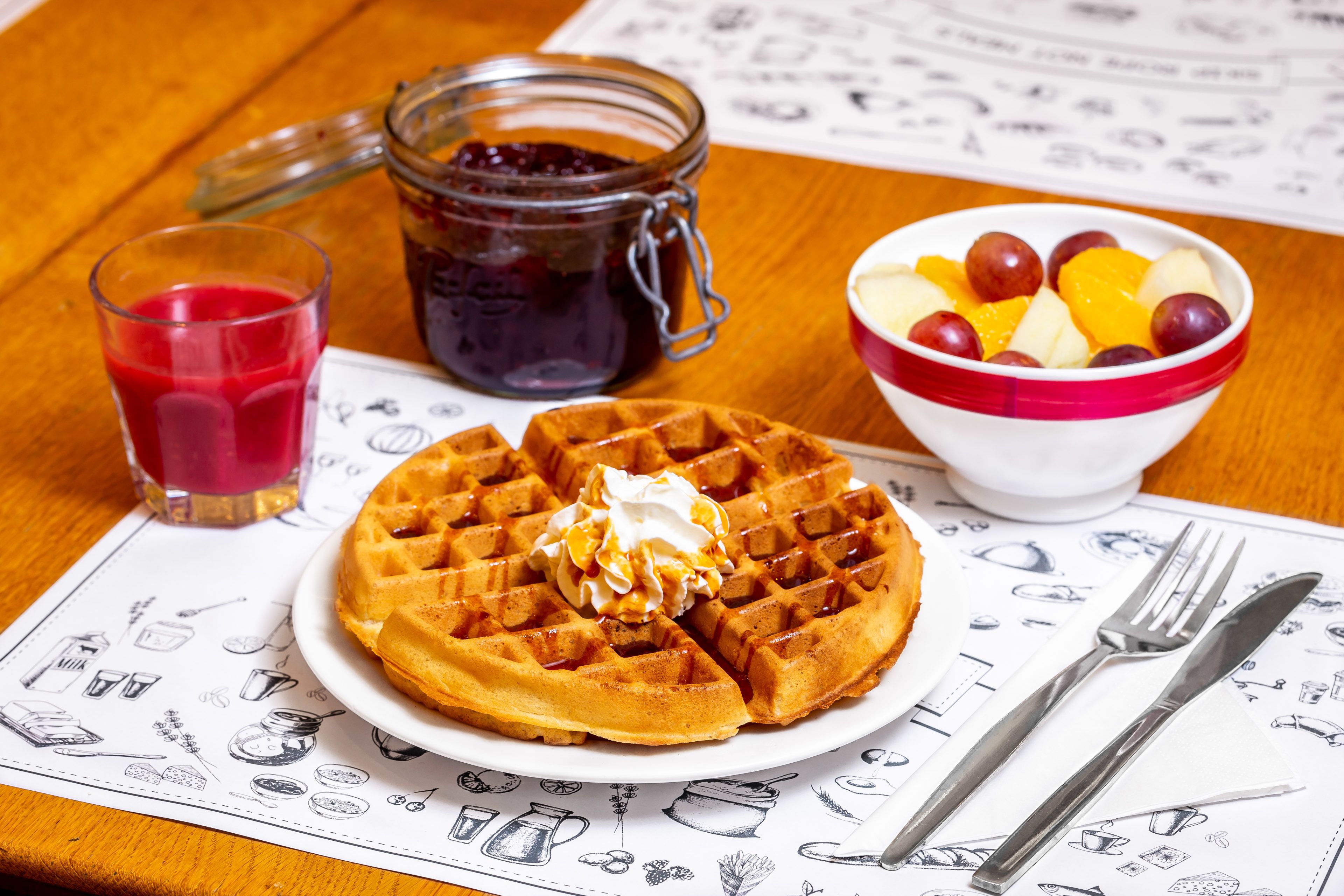 Un plato de waffles con crema batida y jarabe, un bol de fruta, un vaso de jugo rojo y un frasco de mermelada sobre una mesa de madera.