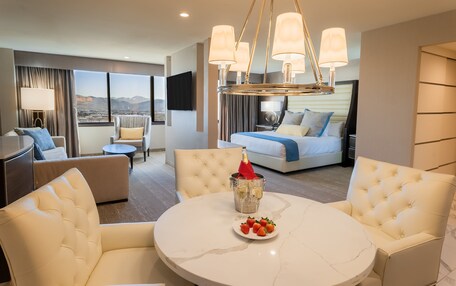 A modern hotel room with a round marble table, plush seating, a minibar, and a large bed with a city view.