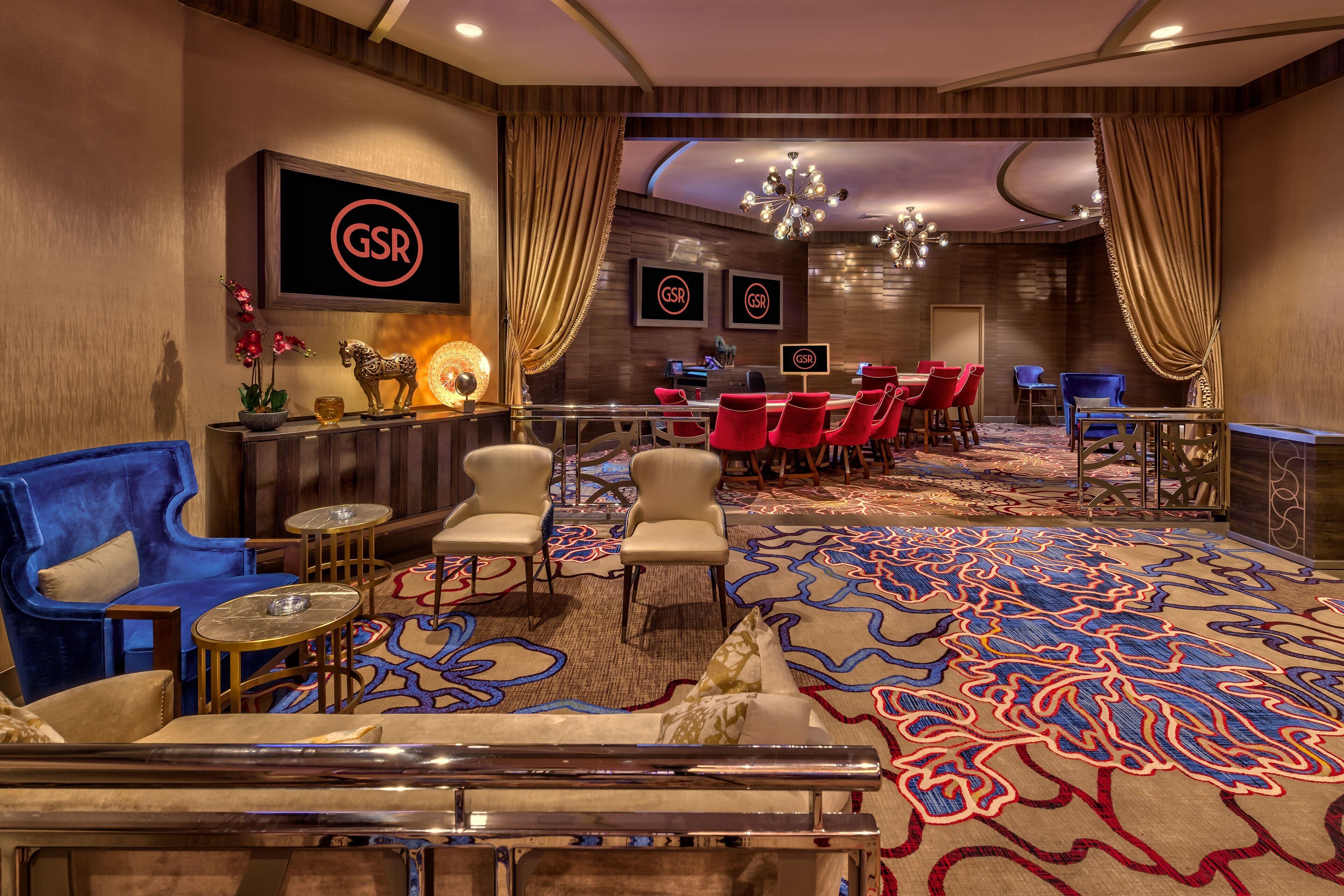 Un hall d’hôtel avec un comptoir d’accueil, des chaises rouges et beiges, un fauteuil bleu et une moquette à motifs.