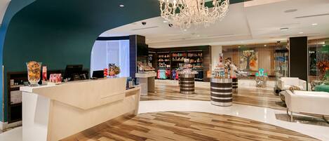 A modern hotel lobby with a reception desk, a chandelier, and a variety of retail displays.