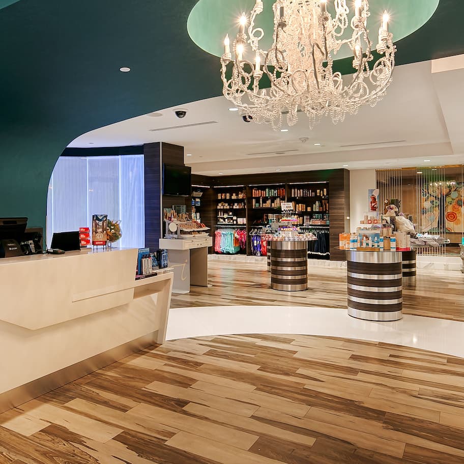 A modern hotel lobby with a reception desk, a chandelier, and a variety of retail displays.