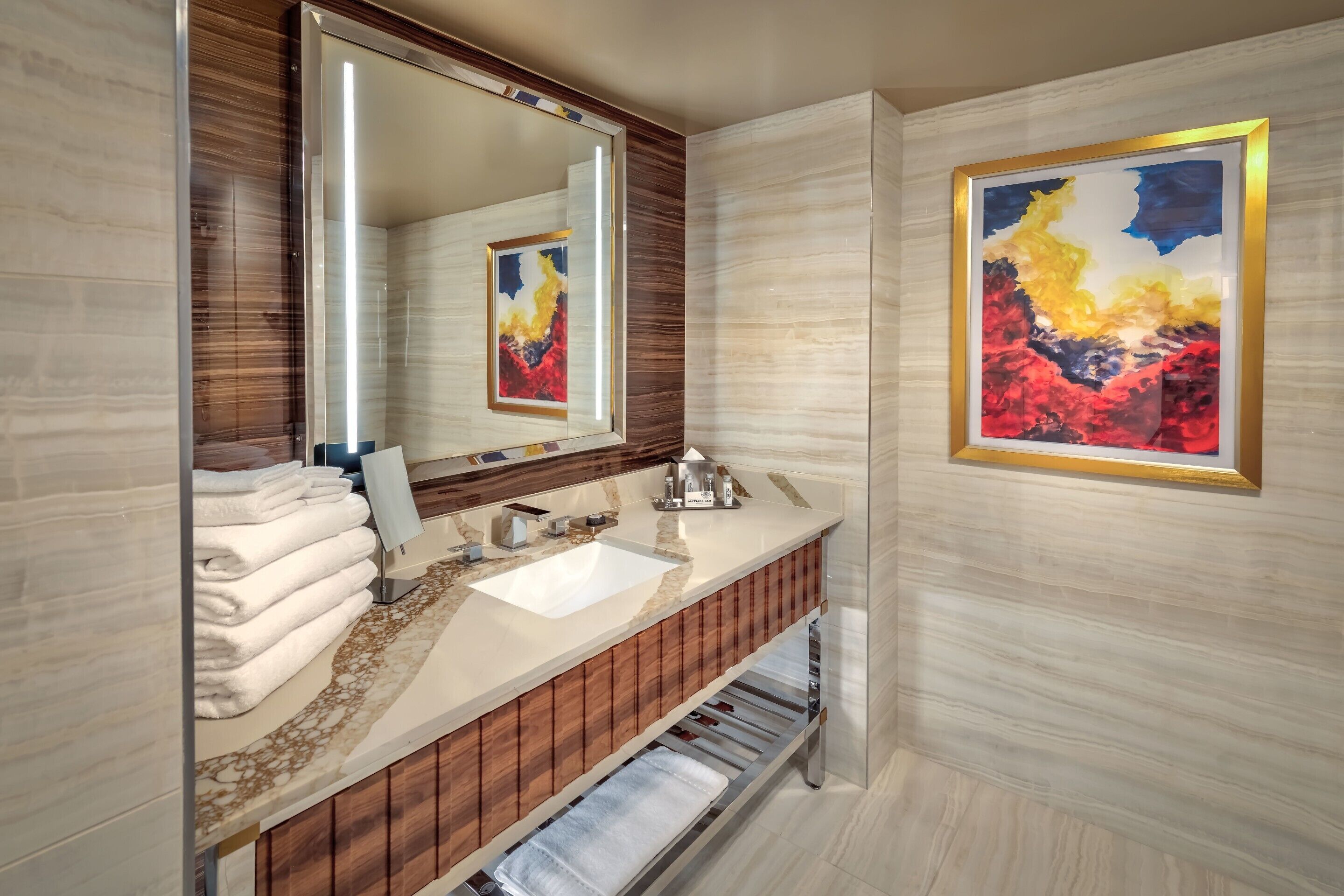 Un baño moderno con un espejo grande, un lavamanos blanco, tocador de madera y un cuadro abstracto enmarcado en la pared.
