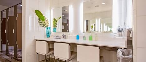 A modern bathroom with a large mirror, white tiles, and a plant in a vase. There are three white chairs and a trash bin.