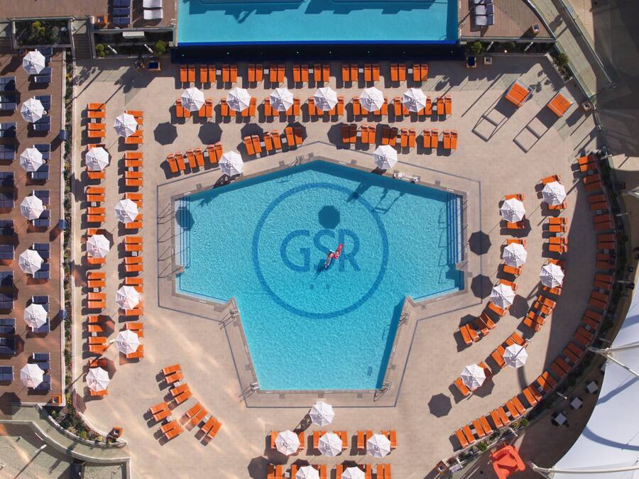 Vista aérea de una zona de piscina con tumbonas anaranjadas y blancas dispuestas alrededor, y un logotipo en el centro.