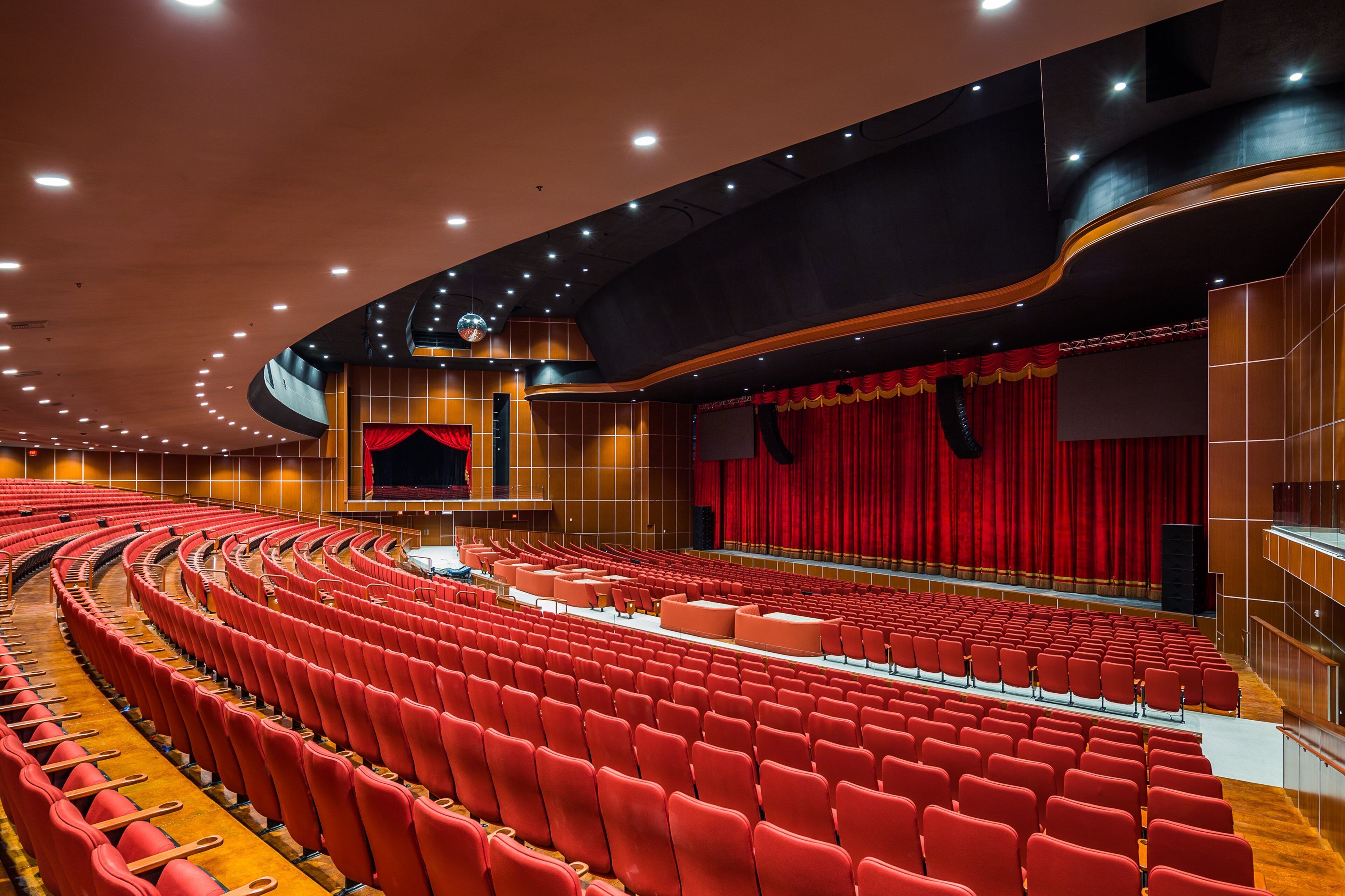 A large auditorium with red seating, a stage with a red curtain, and a modern ceiling design.