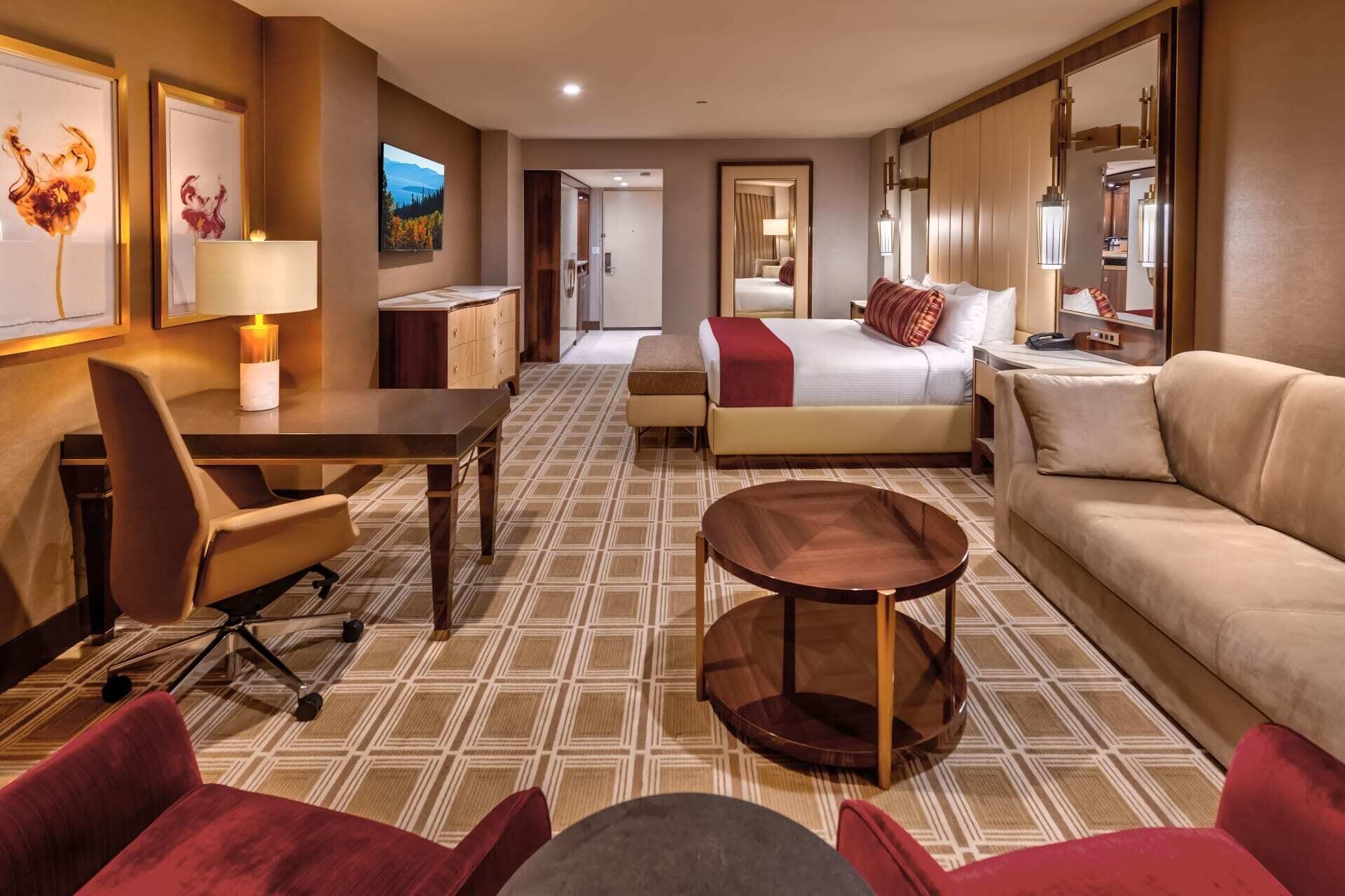 A hotel room with a bed, a sofa, a desk, and a chair. There is a mirror, a lamp, and artwork on the walls.