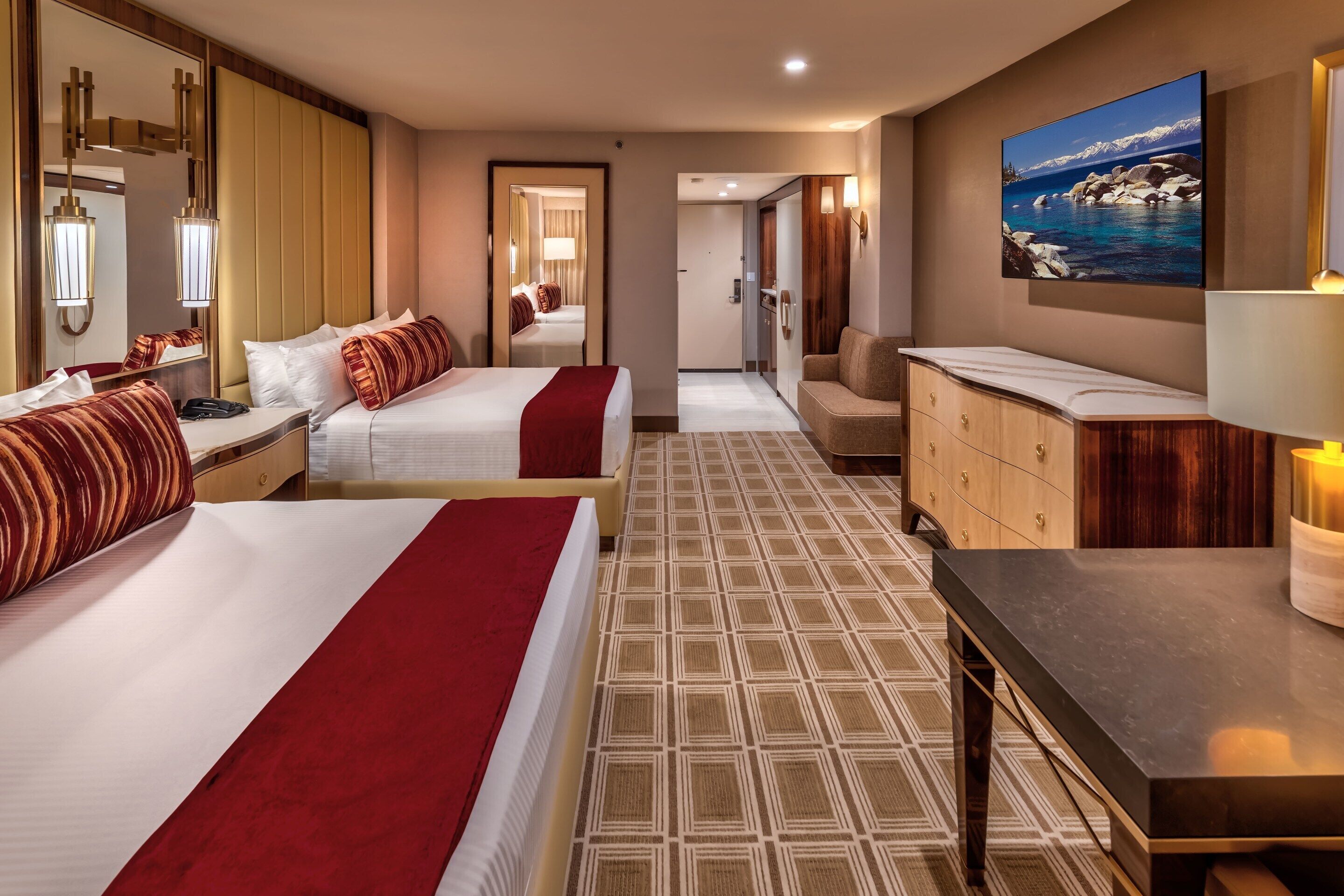 Habitación de hotel con dos camas, una zona de estar, un espejo grande, un cuadro de un paisaje montañoso y un armario de madera.