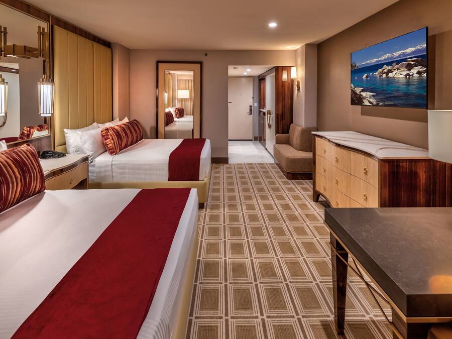 A hotel room with two beds, a sitting area, a large mirror, a painting of a mountain landscape, and a wooden cabinet.