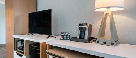 A hotel room with a flat-screen TV, a coffee machine, a lamp, a minibar, and a wooden wardrobe.