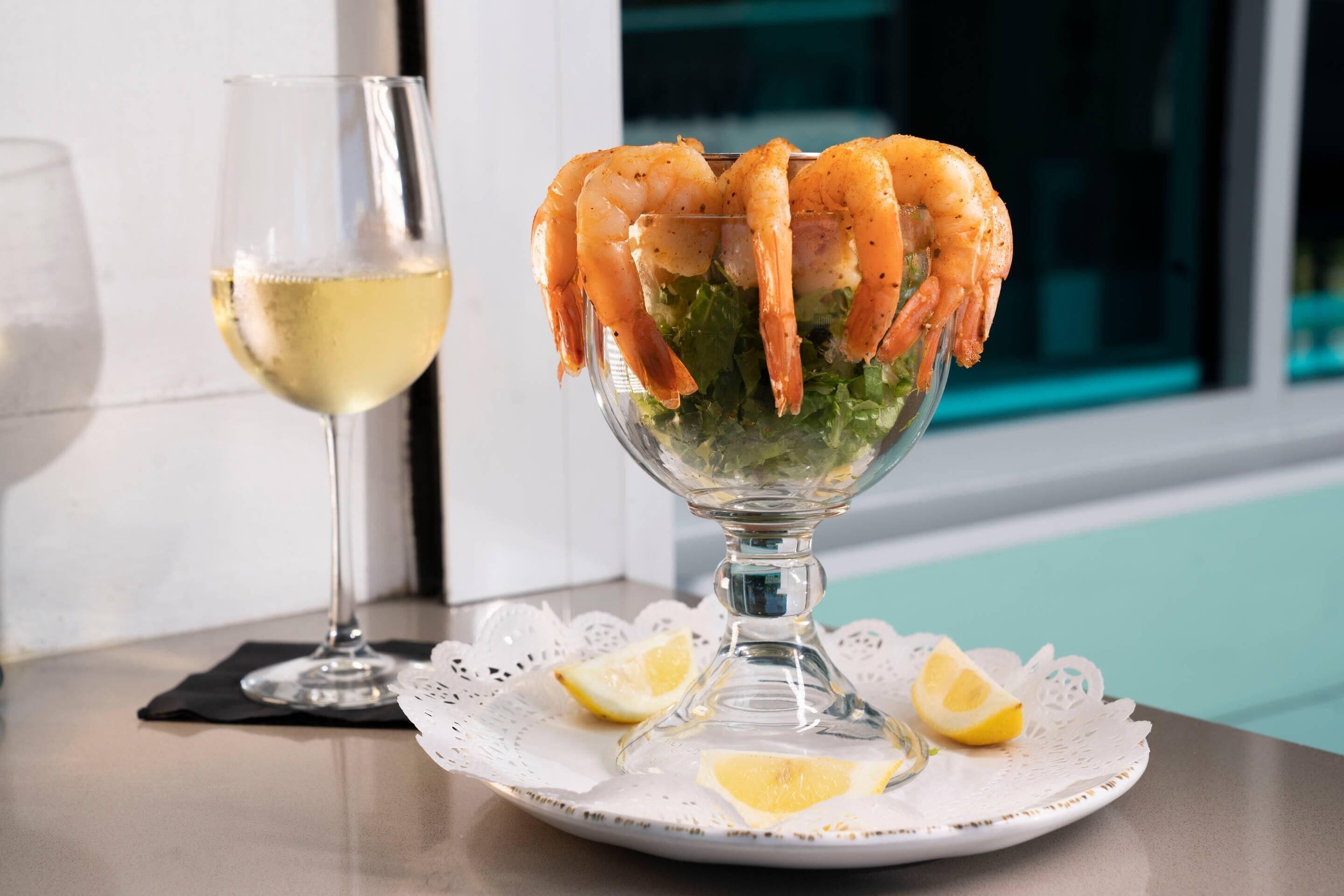 A glass bowl with cooked shrimp on a bed of lettuce, accompanied by a glass of white wine and lemon slices on a white doily.