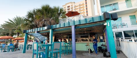 An outdoor seating area with turquoise chairs and umbrellas, a bar area with a person serving, and a building with balconies in the background.