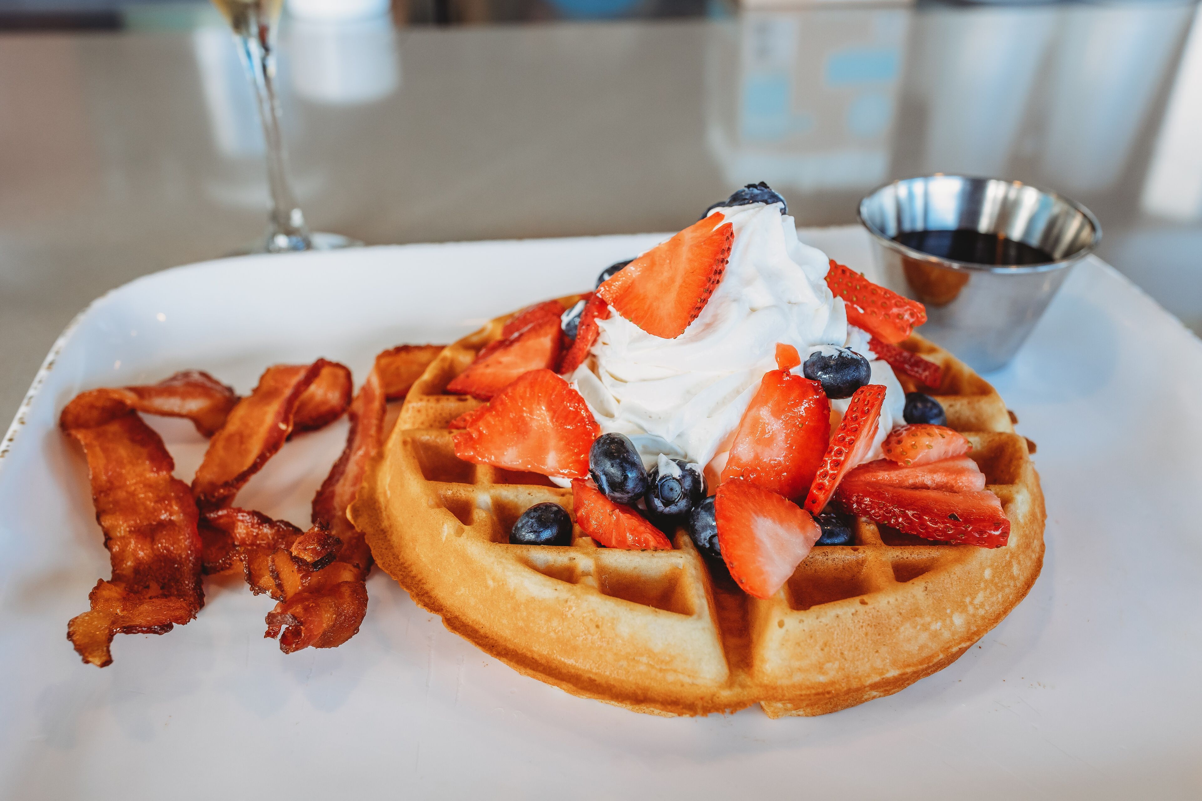 A breakfast plate with a waffle topped with whipped cream, strawberries, blueberries, and bacon strips.