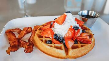 A breakfast plate with a waffle topped with whipped cream, strawberries, blueberries, and bacon strips.