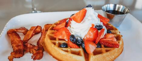 A breakfast plate with a waffle topped with whipped cream, strawberries, blueberries, and bacon strips.